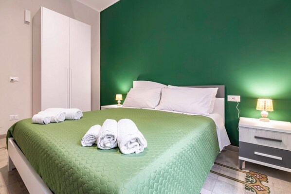 A hotel room with a green bedspread, white pillows, a white wardrobe, a nightstand with a lamp, and a patterned rug.
