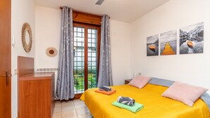 Une chambre avec un lit, un bureau en bois, une vue sur la verdure par la fenêtre et des œuvres d’art mural.