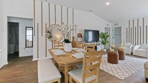 Un salon moderne avec un coin repas, comprenant une table à manger en bois, des chaises à revêtement blanc, un grand miroir et un téléviseur à écran plat.