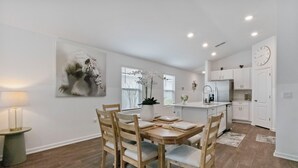 Une cuisine moderne avec des meubles blancs, une table à manger avec des chaises et une grande fenêtre équipée de stores.