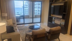 Uma sala de estar com sofá, duas poltronas, uma mesa de centro e televisão. Há uma grande janela com vista para um edifício e para o oceano.