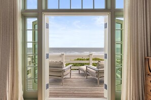 A view from a room with a large window overlooking a wooden deck with two chairs and a table. - Showstopping Oceanfront Home with Pool,  Featured in Southern Living (Kiawah Island)