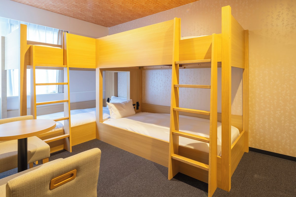 A bunk bed room with a table and chairs, a window with curtains, and a patterned wallpaper.