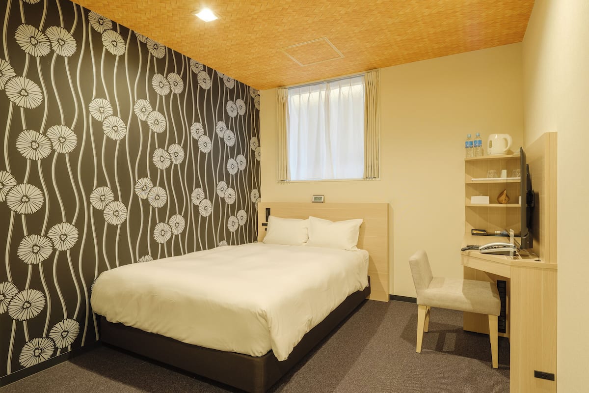 A hotel room with a bed, a desk, a chair, a window with curtains, and a wall with a floral pattern.