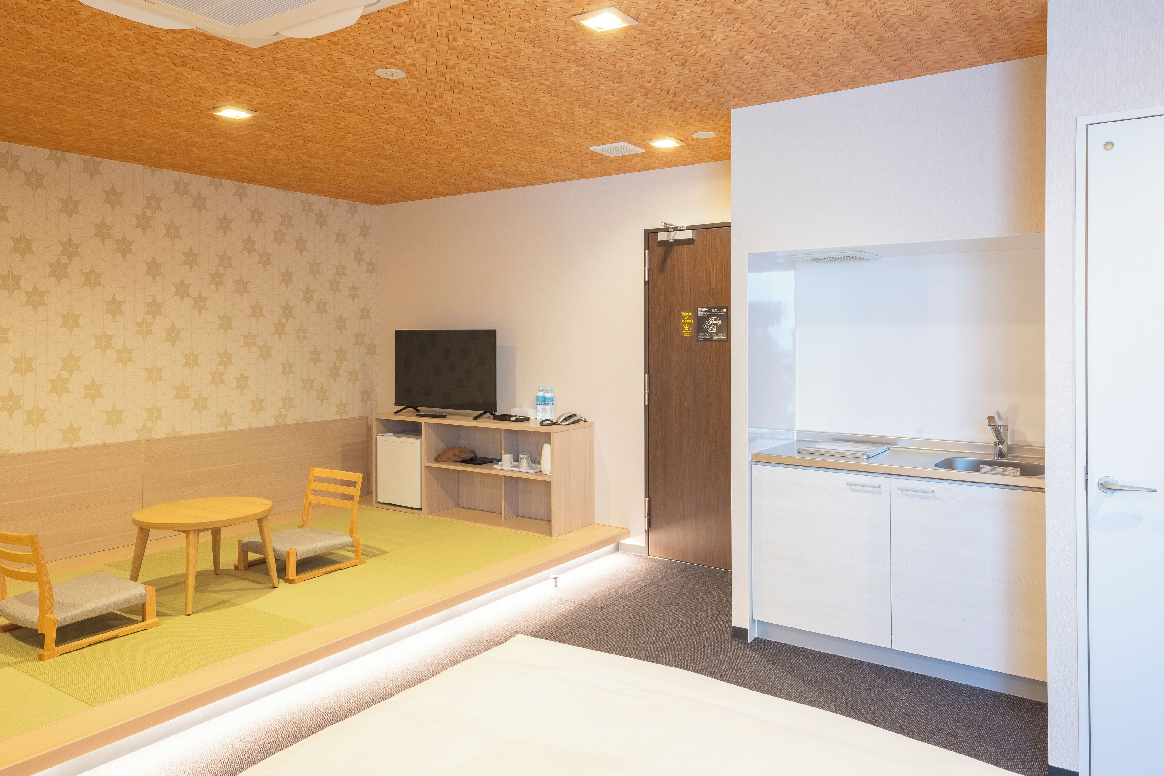 A room with a wooden ceiling, a television on a shelf, a small table with chairs, a kitchen area with a sink, and a door with a sign.