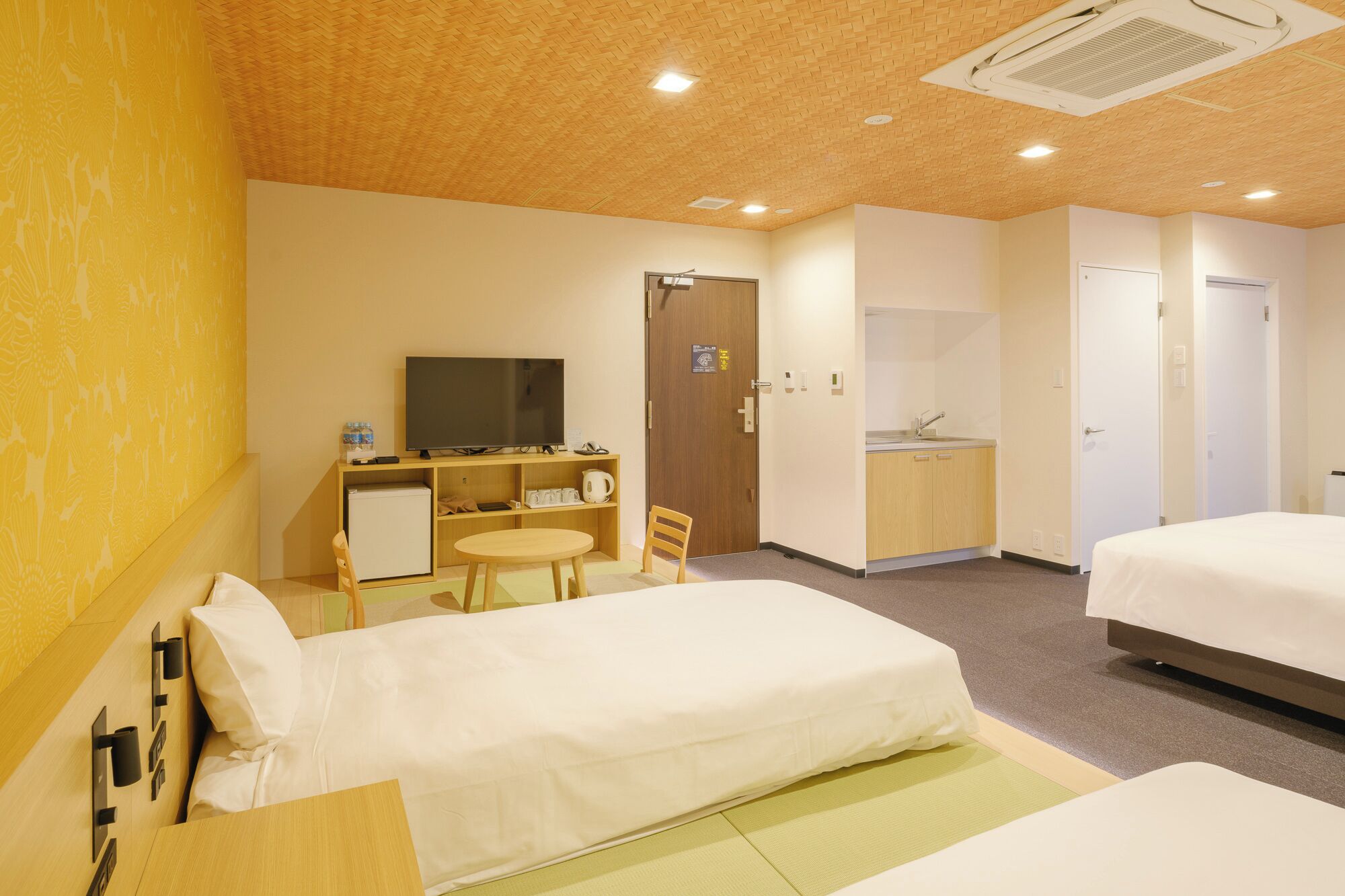A hotel room with two beds, a desk with a TV, a small kitchenette, and a bathroom.