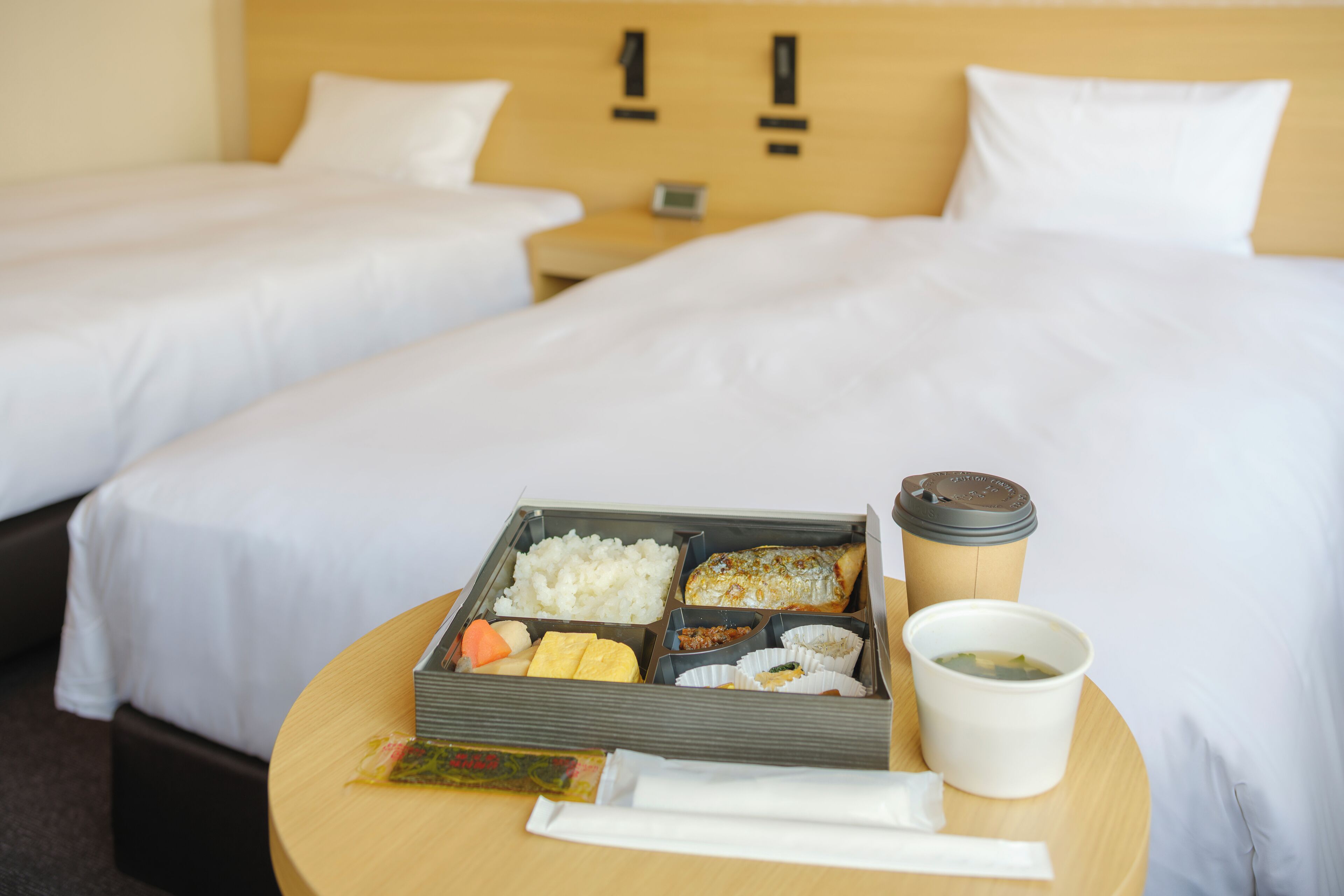 A hotel room with two beds, a bedside table with a tray of food, a coffee cup, and a pillow.