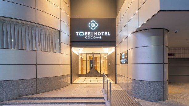 La entrada al Hotel Tosei Cocone, con un diseño moderno, presenta un llamativo letrero iluminado y un frente de vidrio.
