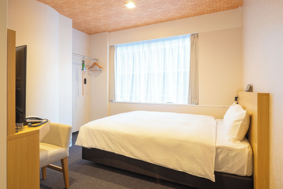 A hotel room with a bed, a desk with a telephone, a chair, and a window with curtains.