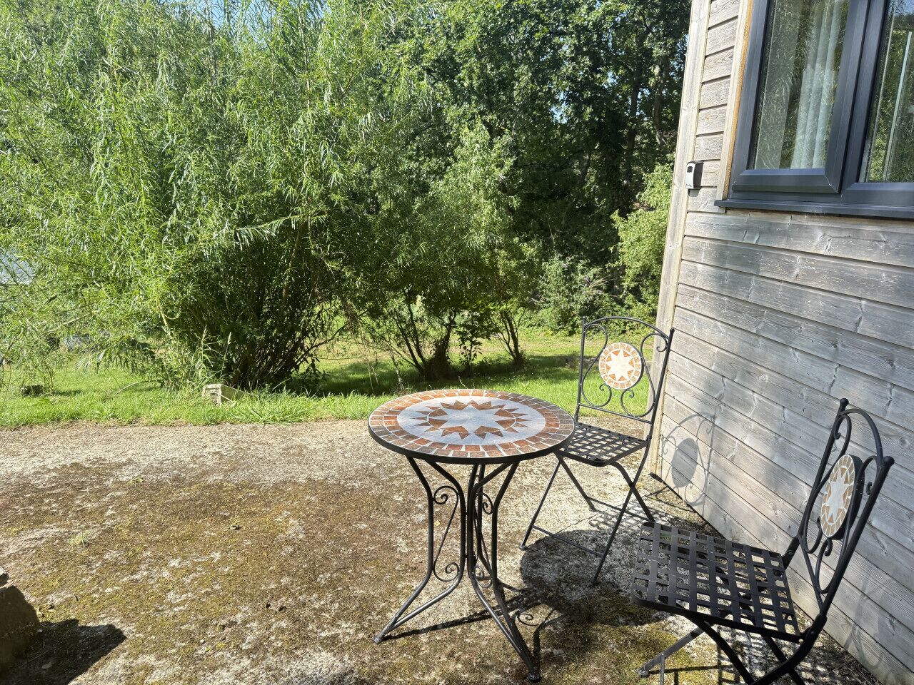 A patio with a round table and two chairs, a small garden, and a wooden building with a window.