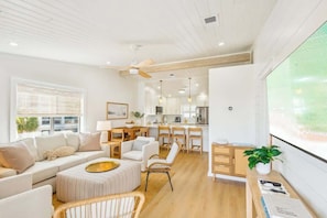 A modern living room with a sofa, ottoman, and chairs, a dining area with a table and chairs, a kitchen with appliances, and a large window.