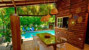 A wooden cabin with a glass table and white chairs, a pool, and tropical plants.