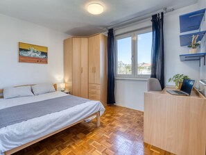 A bedroom with a bed, a wardrobe, a desk with a laptop, a window with curtains, and a painting on the wall.