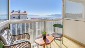 Un balcon avec vue sur les bâtiments et les montagnes, des chaises en osier, une petite table avec une plante et une chaise verte.
