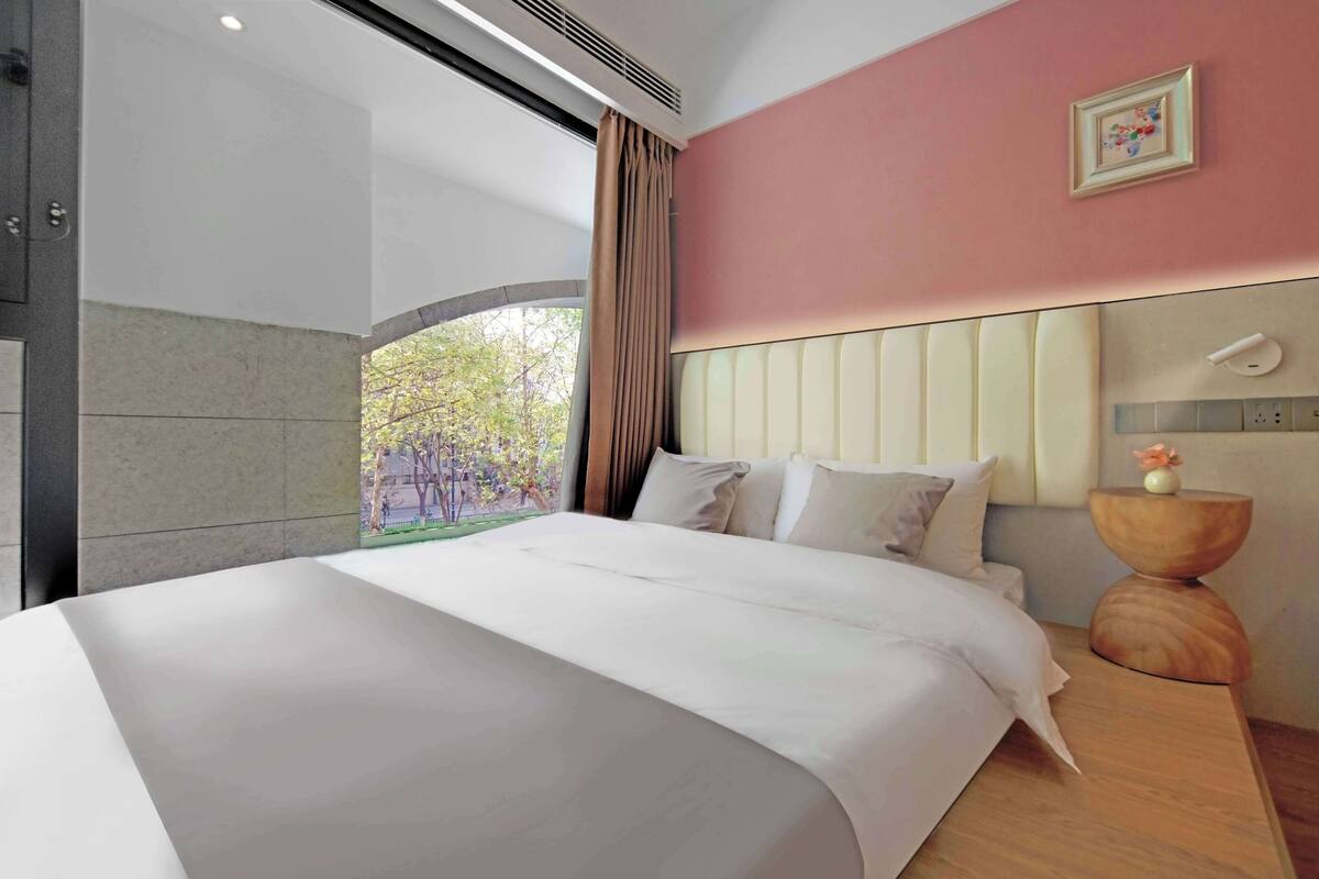 A modern hotel room with a large bed, a wooden nightstand, a wall-mounted light, and a window with curtains.