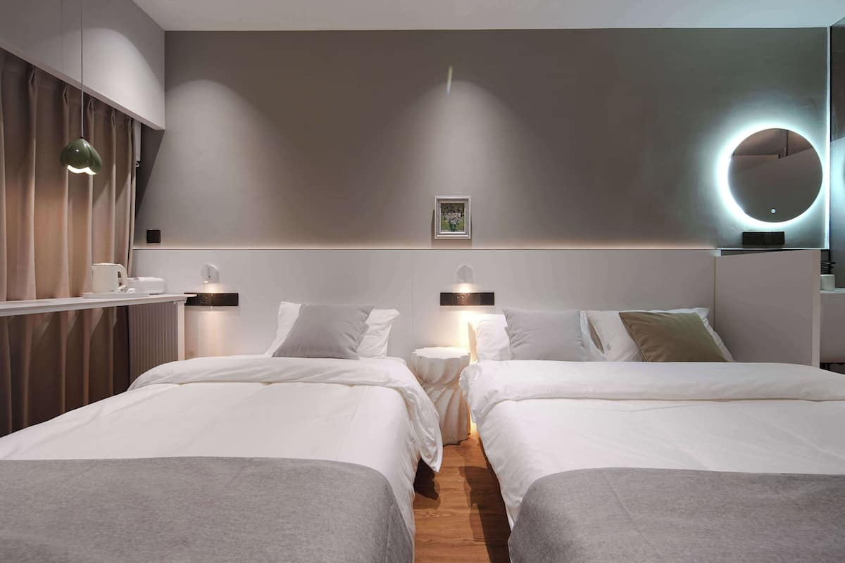A hotel room with two beds, a bedside table, a lamp, a round mirror, and a framed picture on the wall.