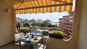 A balcony with a striped awning, a table with chairs, a view of a swimming pool, and buildings in the background. - Discover this charming studio in Santa Margarita, Roses! (Roses)