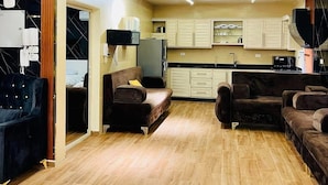 Una sala de estar moderna con un sofá, una mesa de centro y un televisor. Cuenta con una zona de cocina con armarios y un microondas. El ambiente tiene piso de madera e iluminación empotrada en el techo.