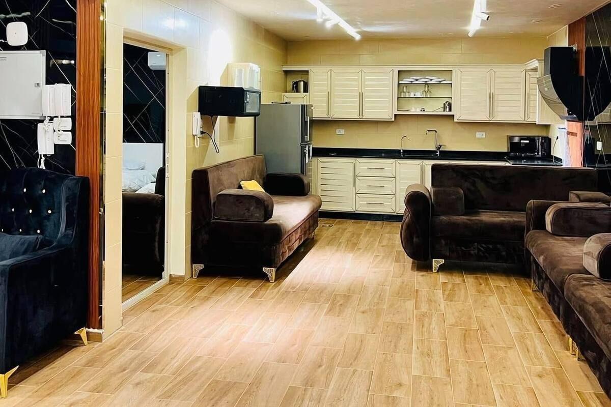 A modern living room with a sofa, a coffee table, and a TV. There is a kitchen area with cabinets and a microwave. The room has wooden flooring and a ceiling with recessed lighting.
