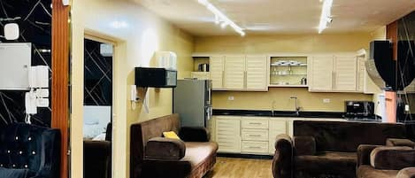 A modern living room with a sofa, a coffee table, and a TV. There is a kitchen area with cabinets and a microwave. The room has wooden flooring and a ceiling with recessed lighting.