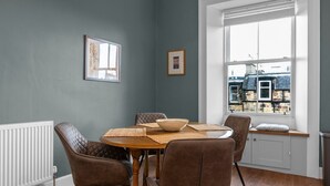 A dining area with a round wooden table, six brown upholstered chairs, a white radiator, and a window with a view of buildings. - GuestReady - Bright & Modern 2BR in Edinburgh (Edinburgh)