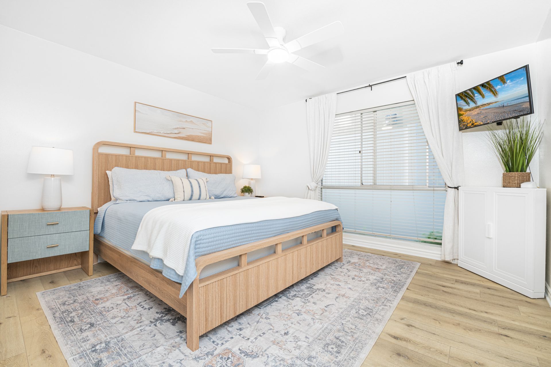 Ein Schlafzimmer mit Bett, Nachttischen, Deckenventilator, einem Fenster mit Jalousien, Fernseher und einer Topfpflanze.