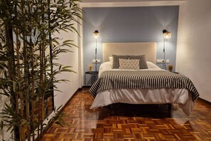 Een slaapkamer met een visgraatvloer, een bed met streepjesbeddengoed, twee bedlampen en een wandlamp.