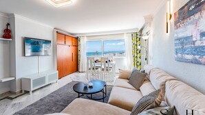 A modern living room with a sofa, a coffee table, and a dining area with chairs. There is a TV mounted on the wall and a wooden door. - Marvelous views, pools, steps to the beach! REF024 (Benalmádena)