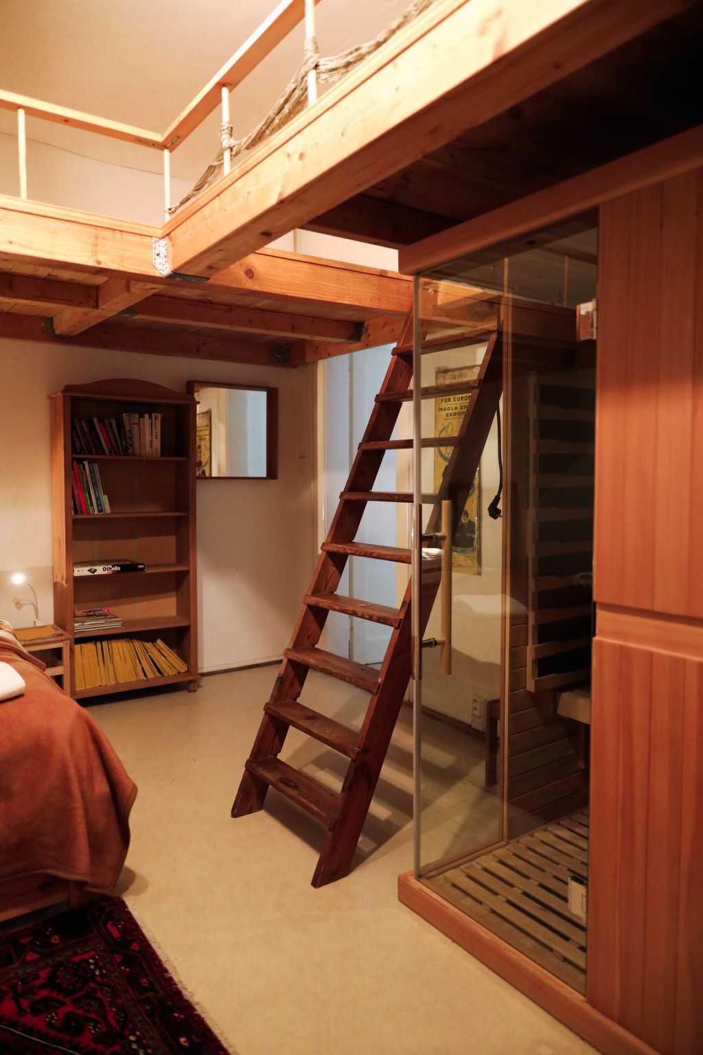 A loft bedroom with a wooden ladder leading to a lofted sleeping area, a bookshelf, and a large window.