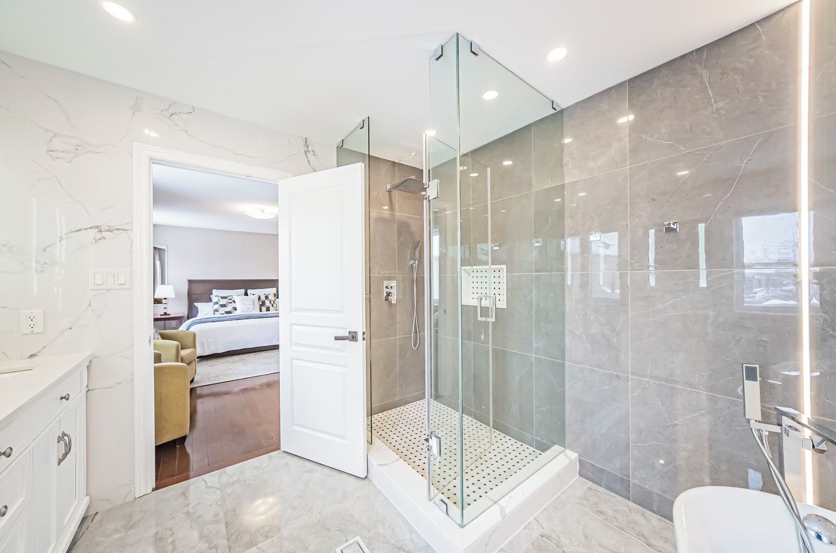 Un baño moderno con ducha de vidrio, paredes de mármol y una habitación visible con cama y mesita de noche.