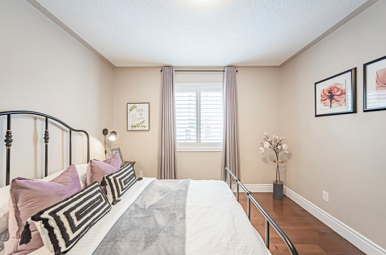 Une chambre à coucher avec un lit, une fenêtre avec des rideaux, une table de chevet avec une lampe et des tableaux encadrés accrochés au mur.