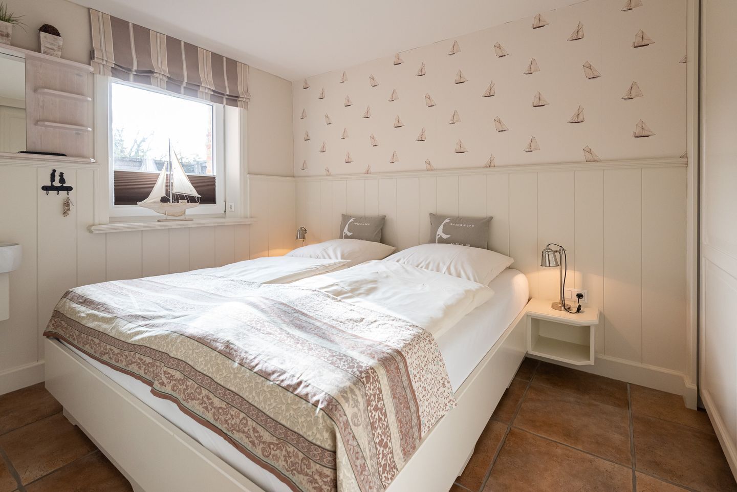 A bedroom with a large bed, a window with curtains, a nightstand with a lamp, and a wall with sailboat wallpaper.