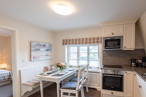 Una cocina compacta con muebles blancos, una mesa de comedor con sillas, una ventana con persianas y un horno empotrado.