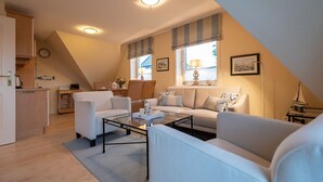 A cozy living room with a sofa, armchairs, and a coffee table. There is a kitchen area with a counter and cabinets, and a dining table with chairs. - Haus Sonnentraum, Whg. 2 - Sun-flooded 2-room apartment on approx. 60 m² for (Sylt)