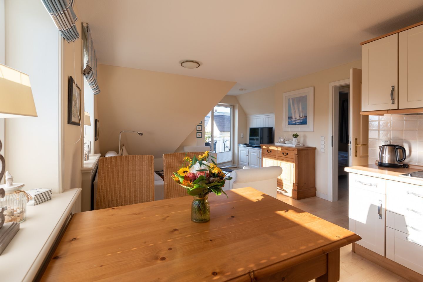 A modern kitchen with white cabinets, a wooden dining table with a vase of flowers, and a view to the outside.