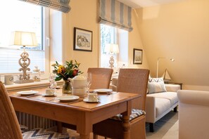 Een eetruimte met een houten tafel gedekt voor vier personen, omringd door rieten stoelen. Op tafel staat een vaas met bloemen en een witte waterkoker. De kamer heeft een gezellige, huiselijke sfeer met een bank en een lamp op de achtergrond.