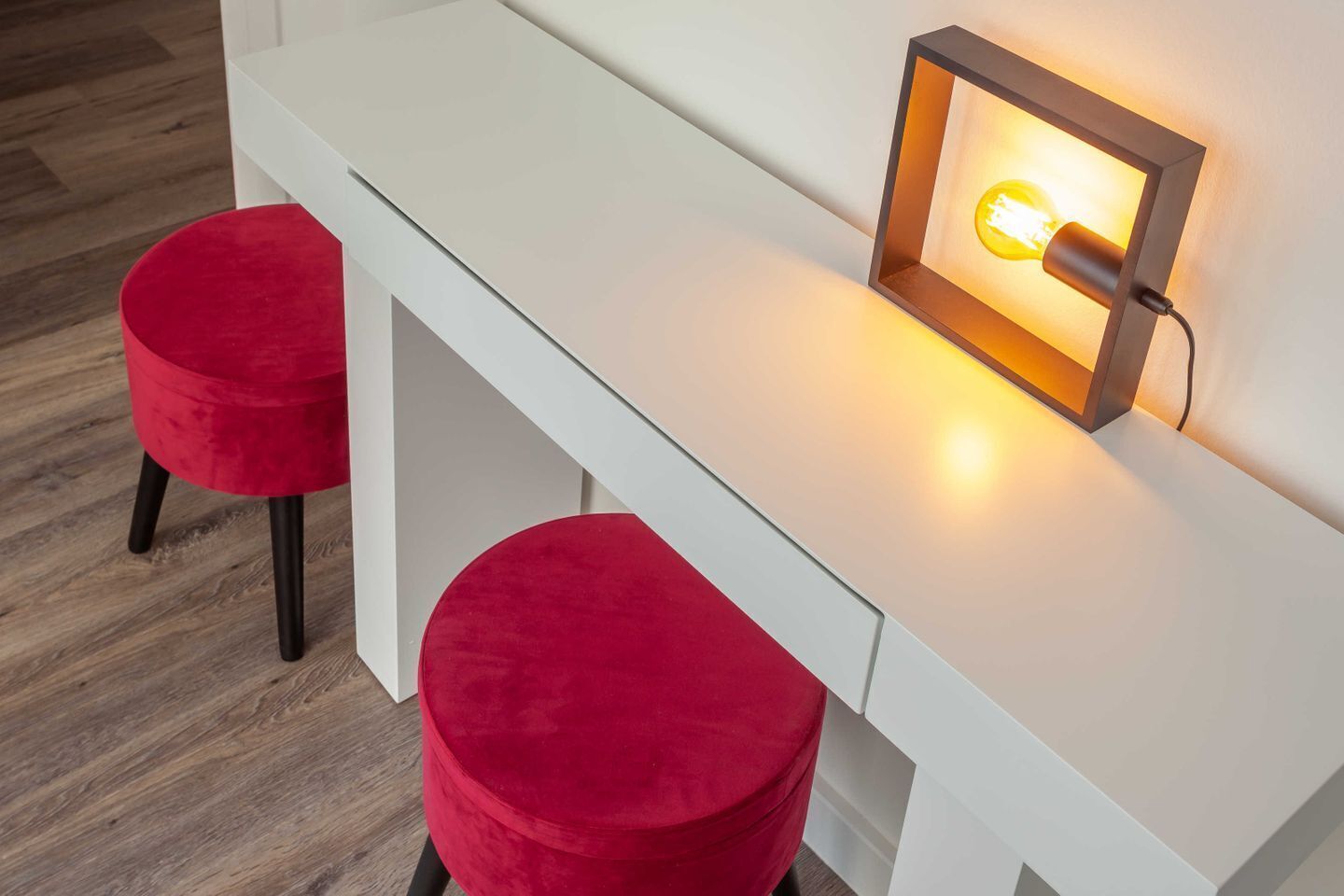 Ein weißer Schreibtisch mit zwei roten Sitzhocker, einer Wandleuchte und einem Holzboden.