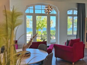 Une pièce avec une table ronde, des canapés rouges et de grandes fenêtres donnant sur un paysage verdoyant et un plan d’eau.