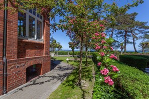 Un bâtiment en briques avec une fenêtre, un chemin et une haie ornée de fleurs roses.