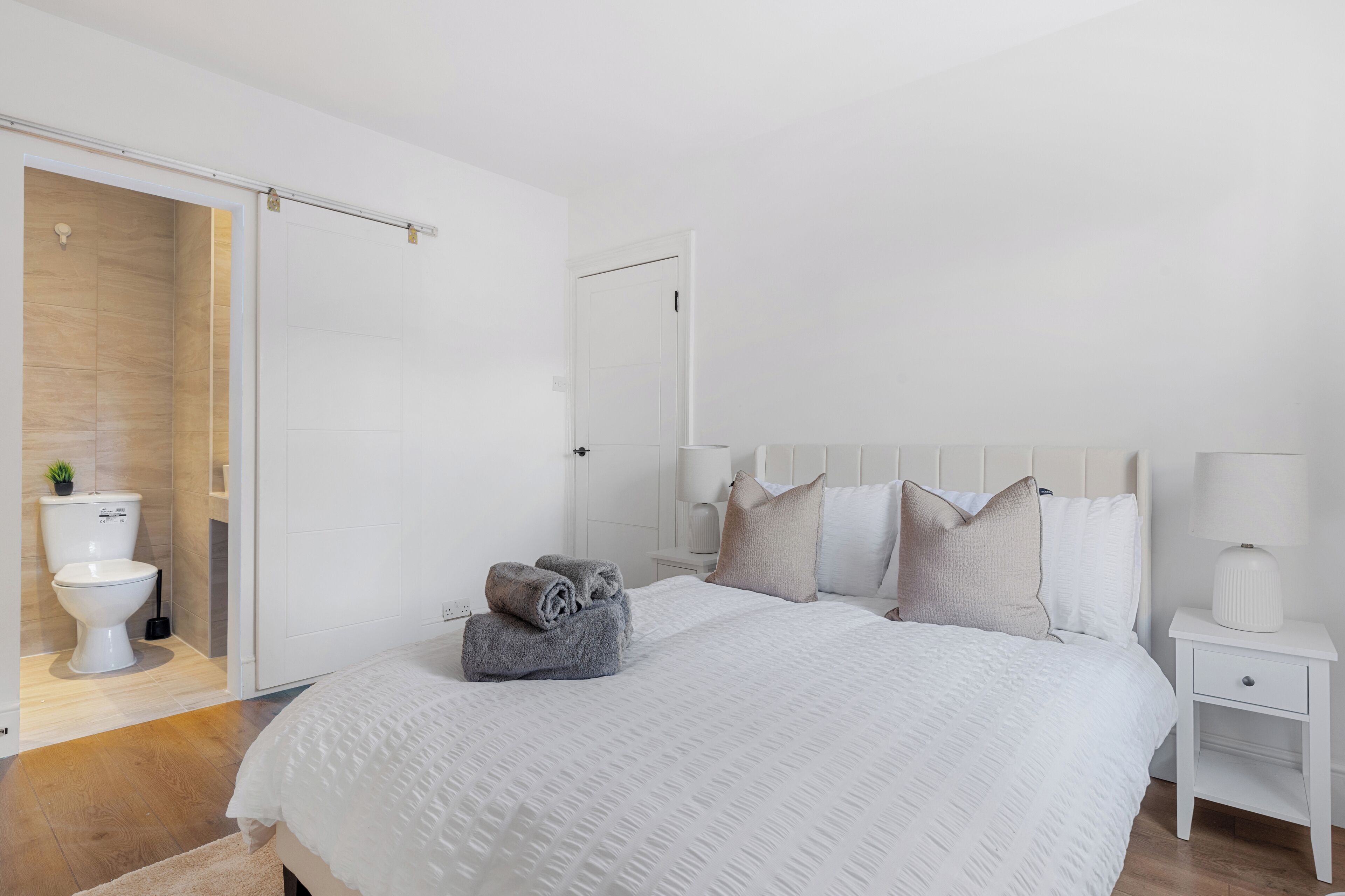Une chambre avec un grand lit, du linge de lit blanc, une table de chevet avec une lampe, et une salle de bain avec des toilettes et un lavabo.
