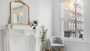 Une pièce avec une cheminée blanche, un miroir doté d’un cadre orné, un globe terrestre, une plante en pot, un fauteuil gris et une vue sur un bâtiment par une grande fenêtre.