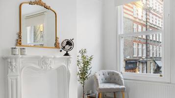 A room with a white fireplace, a mirror with an ornate frame, a globe, a potted plant, a grey armchair, and a view of a building through a large window.