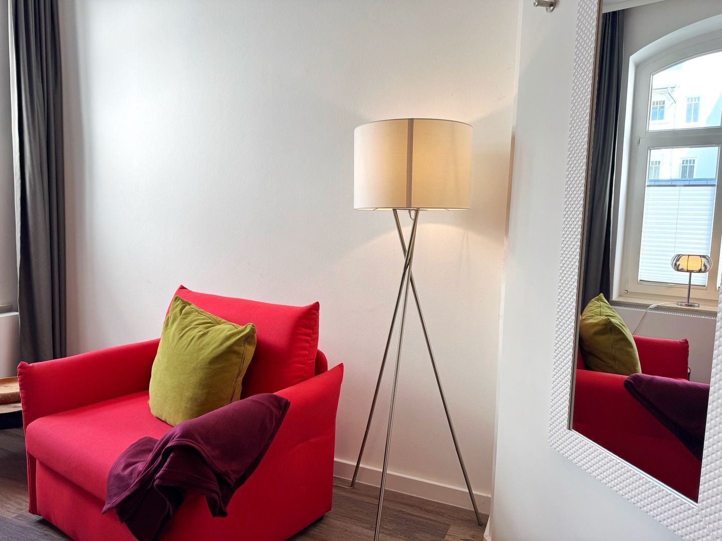 En röd soffa med gröna och lila kuddar, en golvlampa och en spegel som reflekterar en röd stol.