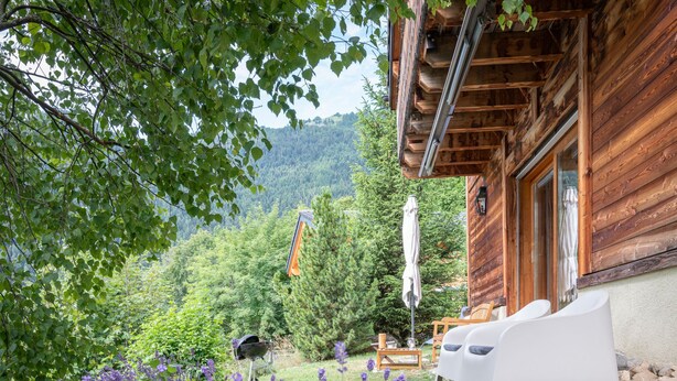 A wooden cabin with a patio area featuring white chairs and a grill. - Dendron by Interhome (Ovronnaz)