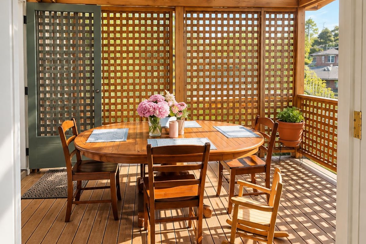 A covered outdoor patio with a wooden table and chairs, a welcome mat, and a view through a wooden lattice fence.