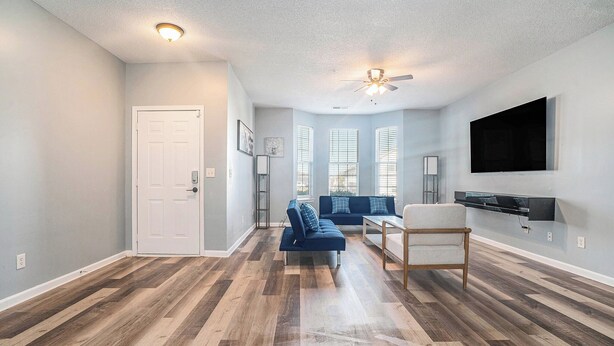A spacious living room with a blue sofa, a wooden floor, a ceiling fan, and a mounted TV.