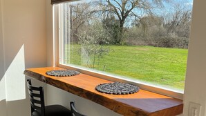 A small kitchen area with a wooden table and two black chairs, a window with a view of a grassy yard and trees, and a built-in oven.