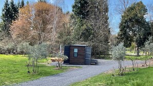 A small wooden cabin with a gravel path leading to it, surrounded by trees and a grassy area.