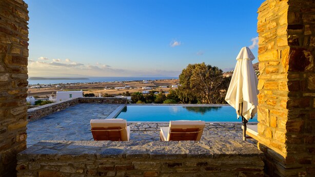 A stone patio with a pool, two lounge chairs, and a view of the sea and distant buildings. - Soho Villa in Paros (Tzanes)