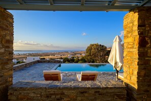 A stone patio with a pool, two lounge chairs, and a view of the sea and distant buildings. - Soho Villa in Paros (Tzanes)